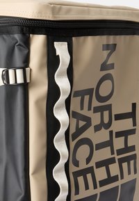 Tansfärgad och svart duffelväska med strukturerad ytfinish, med en vit zigzagrem och stora svarta logodetaljer. Dragkedjestängning i topp.