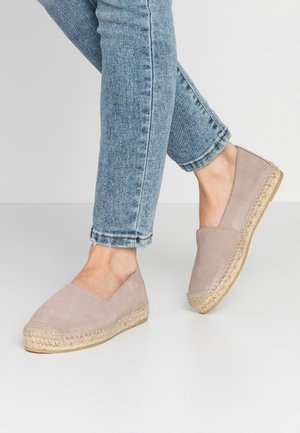Person iført lyseblå denimjeans og beige ruskinds espadrillessko med vævede jutesåler, stående på et hvidt gulv.
