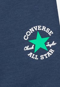 Temno modra tkanina z belim krožnim logotipom, ki ima zeleno zvezdo in besedilo "CONVERSE Chuck Taylor ALL STAR" ukrivljeno okoli logotipa.