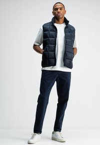 Marineblauer Steppwesten mit gesteppter Optik, Fronttaschen und Stehkragen, getragen über einem weißen T-Shirt und kombiniert mit dunklen Hosen und Turnschuhen.