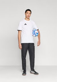 Λευκό αθλητικό πουκάμισο με μαύρες λεπτομέρειες, μαύρο αθλητικό παντελόνι και μαύρα αθλητικά παπούτσια με λευκές ρίγες. Κρατά μια μπλε και κόκκινη μπάλα ποδοσφαίρου.