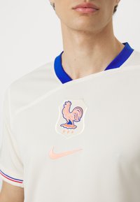 Biała koszulka sportowa z niebieskim kołnierzykiem, z różowym emblematem koguta i logo Nike. Subtelna tekstura, wyszywane akcenty w kolorze czerwonym i niebieskim.