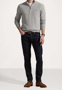 Pull gris à demi-fermeture éclair avec texture tricotée, associé à un jean bleu fonc é et des chaussures en daim marron. Logo discret sur le pull.
