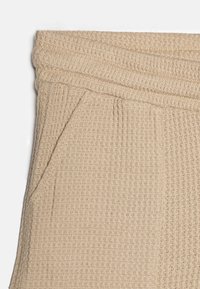 Beige strikkede bukser med et tekstureret mønster, der har en bred talje og sidelommer. Stoffet er strækbart og åndbart.