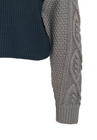 Pull en tricot avec un corps bleu marine et une manche en tricot torsadé gris clair avec des motifs texturés. Les poignets côtelés complètent le design.