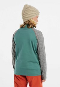 Maglietta a maniche lunghe verde e grigia con colletto alla francese, caratterizzata da una texture liscia e maniche raglan. Indossata con pantaloni arancioni e un berretto beige.