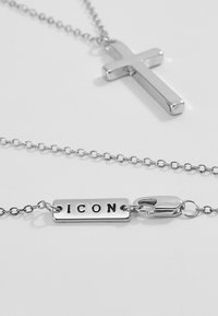 Sølvkjede med et glatt korsanheng og et rektangulært tag med inskripsjonen "ICON." Teksturert kjede med karabinlås.