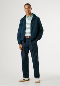 Chaqueta con capucha azul marino sobre una polo a rayas verdes y blancas, con pantalones caqui y zapatillas blancas con detalles en azul marino.