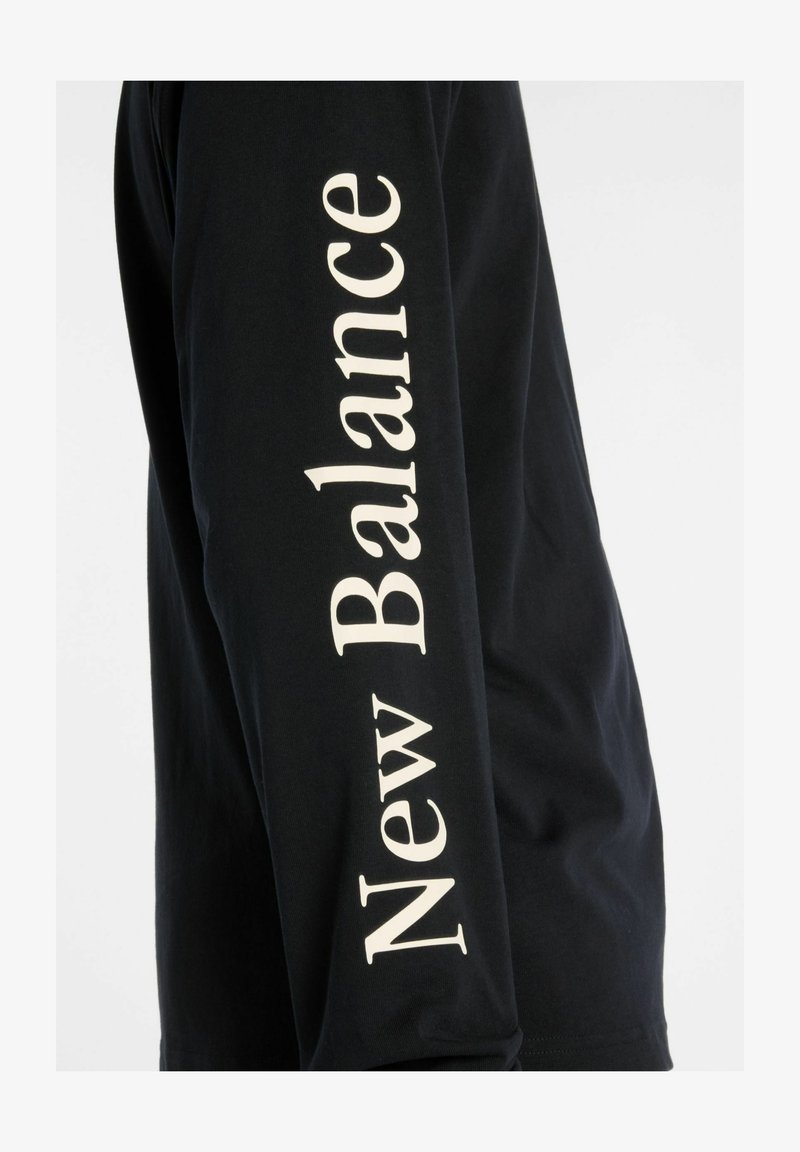 Black long-sleeve t-shirt made of cotton, featuring "New Balance" logo in large white font along the arm. Smooth texture and standard fit.