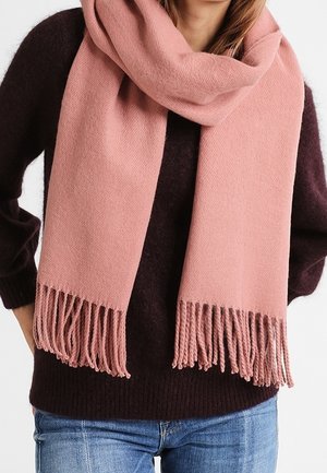 Personne portant une épaisse écharpe rose clair à franges sur un pull marron foncé et un jean bleu, debout les mains sur les hanches.