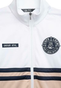 Balta užtrauktuku užsegama striukė su juodais ir smėliniais horizontaliais dryžiais, apvaliu logotipu ir "UNFAIR ATHL." etikete krūtinėje.