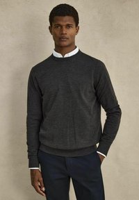 Mörkgrå stickad tröja med rund hals och ribbade ärmar, bärs över en vit skjorta med krage, ihop med marinblå byxor. Enkelt mönster.