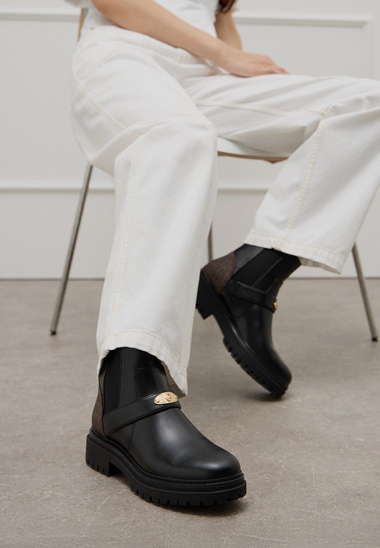 Czarne skórzane botki za kostkę z błyszczącym wykończeniem, brązowe zamszowe akcenty, złote detale oraz solidna gumowa podeszwa, stylizowane z białymi spodniami.