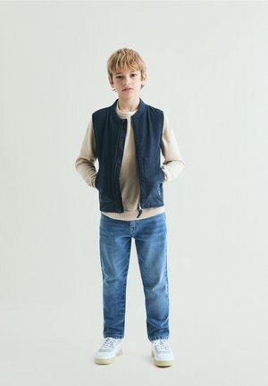 Niño rubio de pie con las manos en los bolsillos, vestido con suéter beige, chaleco azul marino, vaqueros azules y zapatillas blancas sobre un fondo claro y liso.