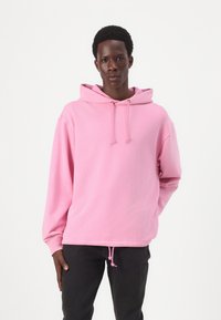 Hoodie rose en tissu doux avec capuche à cordon de serrage, poignets côtelés et coupe décontractée, assortie à un jean foncé. Texture lisse dans l'ensemble.