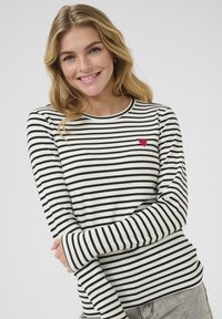 Camicia a maniche lunghe a righe in bianco e nero con una toppa a forma di cuore rosso sul lato sinistro del petto; tessuto morbido con collo tondo.