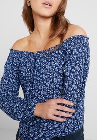 Blue floral off-the-shoulder blouse featuring a gathered neckline and long sleeves, with small white and light blue flower patterns.