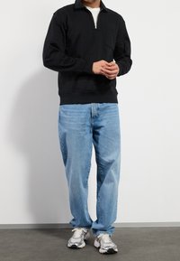 Hombre vestido con un pulóver negro de media cremallera, jeans holgados de color azul claro y zapatillas plateadas, de pie contra una pared blanca con las manos entrelazadas.