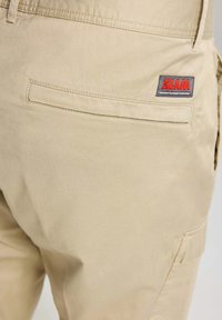 Beige cargobroek met een gladde textuur, voorzien van een achterzak en een logo patch in rood en grijs. Eenvoudig ontwerp met praktische details.