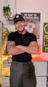 Melns īsroku polo krekls, pelēkas bikses, sakrustotas rokas. Telpā ar augiem, sporta apaviem un plakātiem fonā.