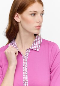 Rosa Polohemd mit einem gemusterten Kragen, der wei�ße und lila Punkte aufweist; glatte Textur und drei Knöpfe an der Vorderseite.