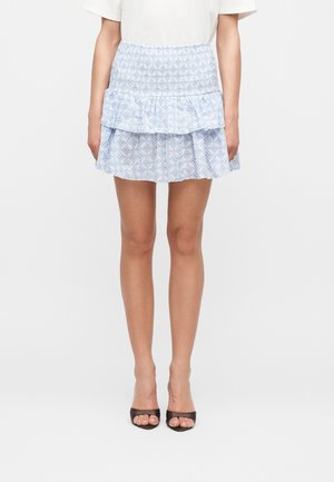 Femme portant un haut blanc avec une mini-jupe à volants à motif bleu clair et des talons ouverts noirs, debout devant un fond uni.