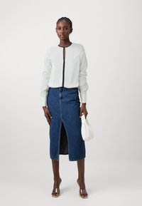 Bluză de culoare verde deschis, cu un detaliu de fermoar negru, purtată cu o fustă din denim cu talie înaltă, care are o deschidere frontală, și sandale cu toc transparent.
