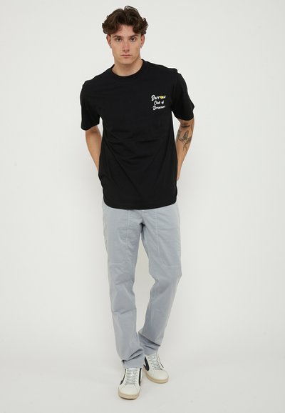 Young man with tattoo wearing black "Barrow Club of Dreamers" t-shirt, light gray pants, and white sneakers standing against white background.
