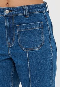 Κοντινό πλάνο της μέσης με ψηλόμεσα μπλε τζιν παντελόνια, με πίσω τσέπη, ασημένιο κουμπί και θηλιές ζώνης σε ανοιχτό φόντο.