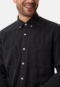 Chemise noire à boutons en coton avec un col, deux poches poitrine et des boutons blancs contrastants, présentant une texture lisse et un design décontracté.