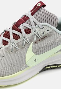 Primer plano de una zapatilla de trail running Nike en gris y burdeos con suela amarilla pálida, cordones gris claro y parte superior de tela de malla transpirable.