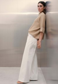 Frau lehnt an einer reflektierenden Wand und trägt einen beige gerippten Pullover, weiße Hosen mit weitem Bein und offene Sandalen in einer neutralen Umgebung.