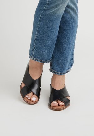 Stopy w niebieskich dżinsach noszące czarne skórzane sandały z szerokimi krzyżującymi się paskami i brązowymi podeszwami na jednolitym białym tle.