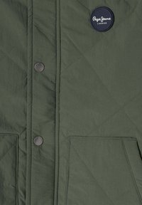 Dunkelgrüne gesteppten Jacke mit silbernen Druckknöpfen, einem runden dunklen Aufnäher mit "Pepe Jeans London" und zwei Fronttaschen.