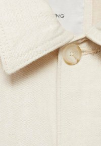 Tessuto di camicia in cotone beige con colletto a punta, caratterizzato da un bottone tondo e testurizzato e dettagli di cucitura visibili.