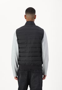 Black puffer vest with a high collar, quilted design, smooth texture, and elastic hem. Worn over a light gray long-sleeve shirt.