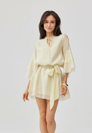 Žena s dlouhými tmavými vlasy v světle krémových šatech s volánky, zvonovými rukávy a páskem v pase, stojící před jednobarevným světlým pozadím.