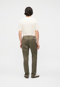 Άνδρας με σκούρα κυματιστά μαλλιά, φοράει κρεμ πουκάμισο με κοντά μανίκια, ελαιδί παντελόνι και μπεζ παπούτσια, στέκεται με την πλάτη σε απλό φόντο.