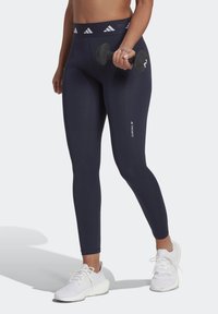 Leggings alta vita blu navy con cintura testurizzata e logo riflettente; il modello tiene un manubrio, indossando scarpe sportive bianche.