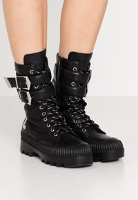 Botines de cuero negro con detalles de hebilla plateada, punta redonda, suela de caucho texturizada y cordones contrastantes en negro y blanco.