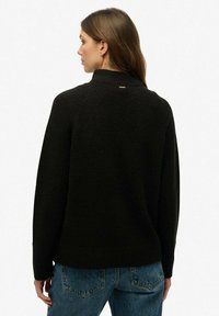 Svart stickad tröja med hög krage och ribbade manschetter, med en subtil guldaccent på baksidan. Kombineras med blå denimjeans.