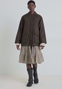 Veste marron mi-longue avec deux poches avant ; jupe écossaise à plusieurs couches ; bottes en cuir noires montantes avec boucles ; fond clair.
