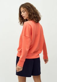 Felpa corallo con fit rilassato, maniche lunghe e polsini a costine, abbinata a pantaloni corti blu navy. Texture liscia e design semplice.