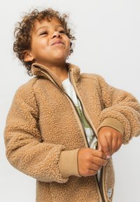 Νεαρό παιδί με σγουρά μαλλιά, που φοράει μπεζ χνουδωτό μπουφάν με φερμουάρ, χαμογελά και κοιτάζει ελαφρώς προς τα πάνω, με φόντο ένα απλό ανοιχτόχρωμο φόντο.