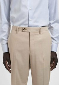 Pantaloni beige su misura con una texture liscia, dotati di chiusura a bottone e passanti per la cintura. Abbinati a una camicia colletto azzurro chiaro.
