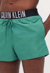Zelene kupaće gaće s crnim elastičnim pojasom s oznakom "CALVIN KLEIN", zatvaranjem na vezivanje i glatkim, laganim materijalom.