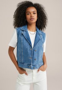 Denimväst med strukturerad krage, dubbelknäppning med metalldetaljer och ljusblå tvätt, bärs över en vit skjorta.