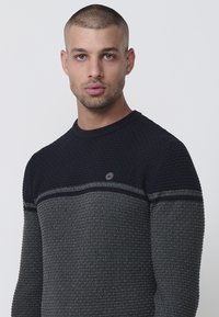 Suéter de textura oscura con un diseño estampado en colores azul marino y gris, cuello redondo y un pequeño logotipo en el pecho.