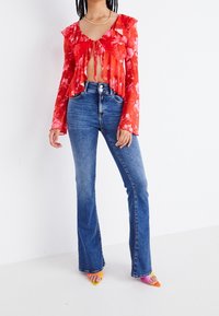 Mujer que lleva unos pantalones de campana de tiro alto de color azul, una blusa corta de manga larga con estampado floral rojo y un lazo en la parte delantera, y tacones de punta coloridos, de pie sobre un suelo blanco.