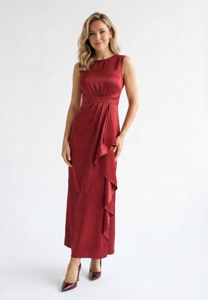 Femme en robe de soirée en satin rouge sans manches avec un détail de volants sur le côté et des talons assortis, debout sur un fond blanc.